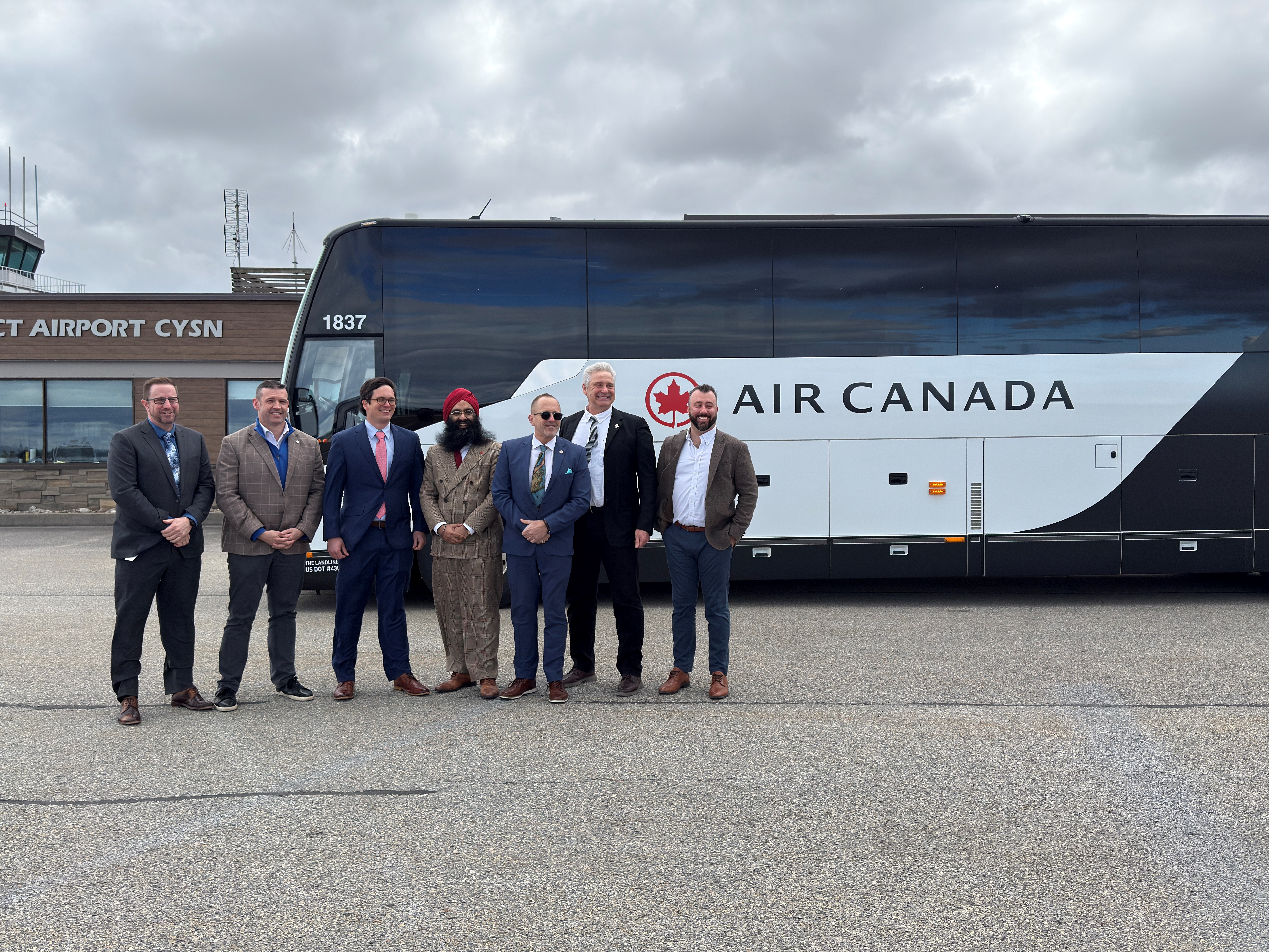 Seven people stand in front of a bus with an Air Canada decal on it, parked in front of the Niagara District Airport terminal.
