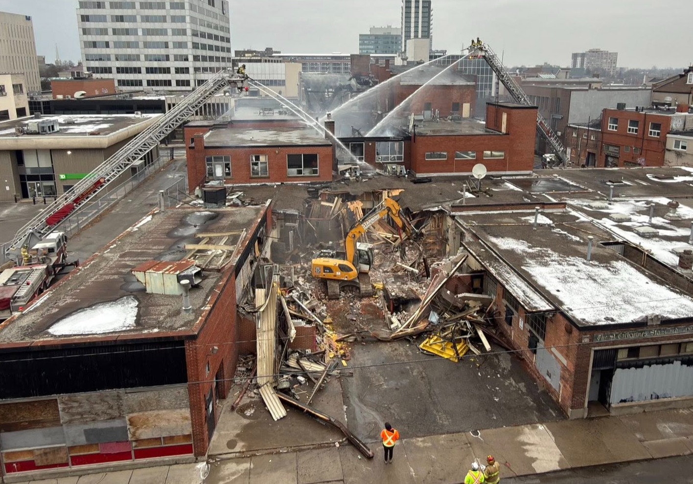 Fire crews using an aerial truck hose down a large building after a fire, as an excavator tears through the bottom of the building.