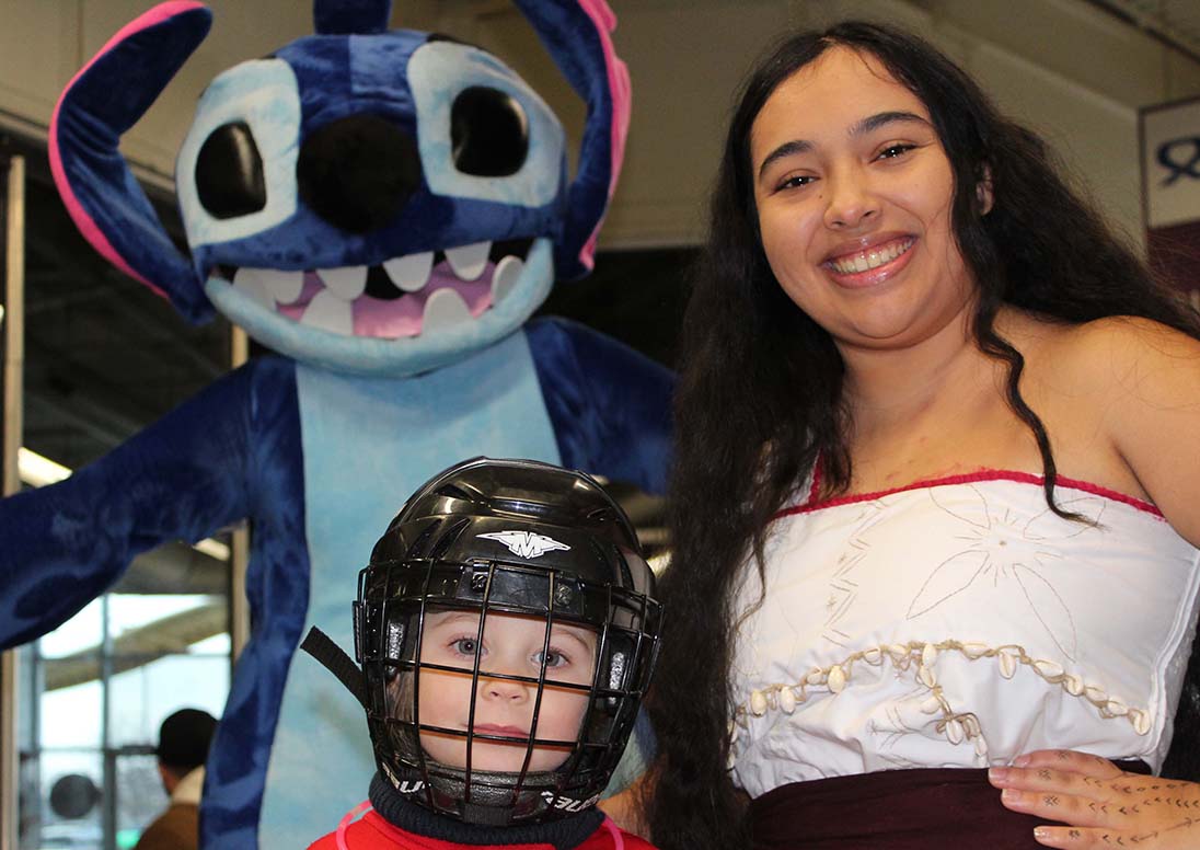 A child in a hockey helmet takes a break from the ice to pose for a photo with Stitch and Moana during the New Year's Eve Public Skate in 2024.