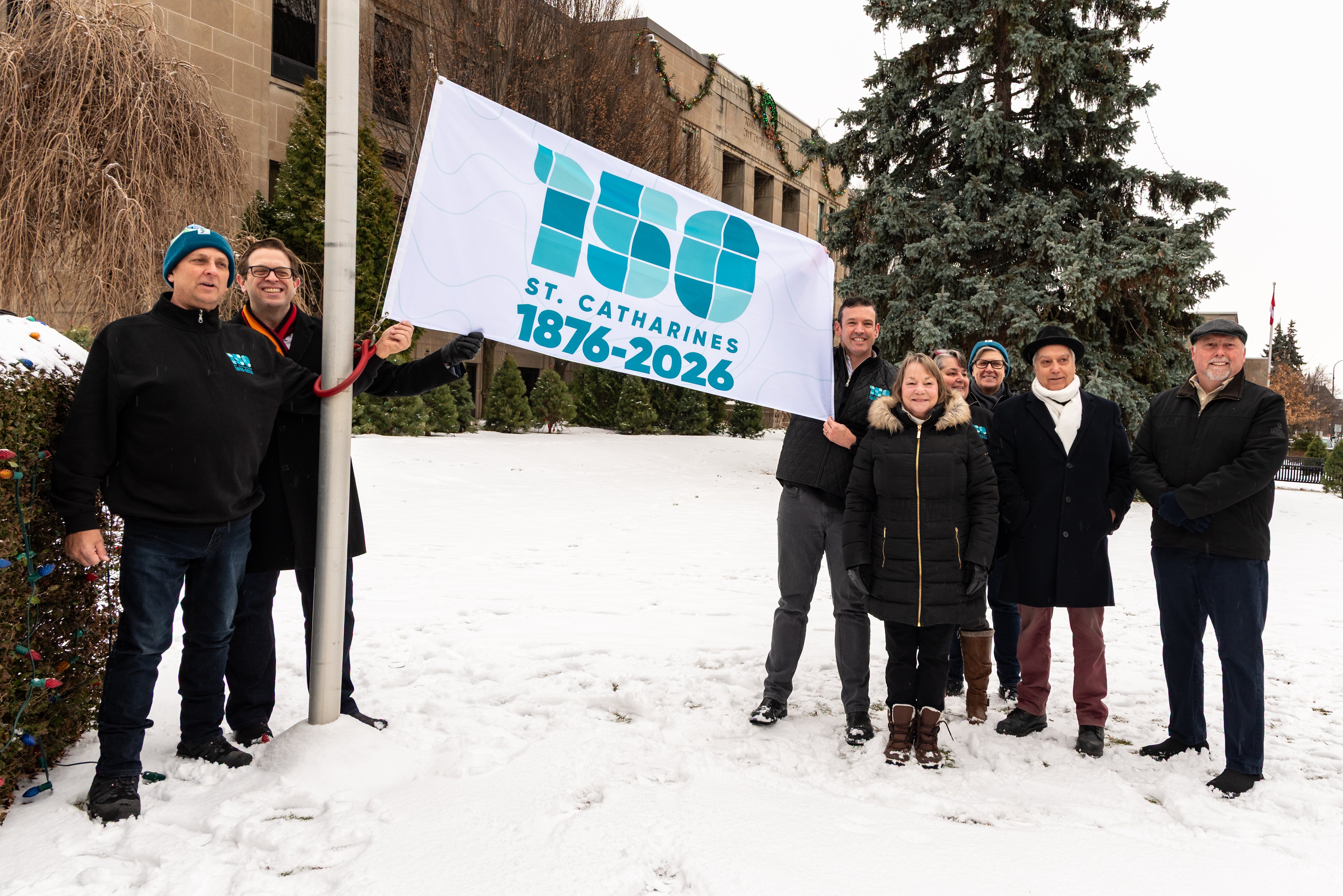 People hold a flag with the number 150 and the name St. Catharines and the dates 1876 to 2026 on the flag.