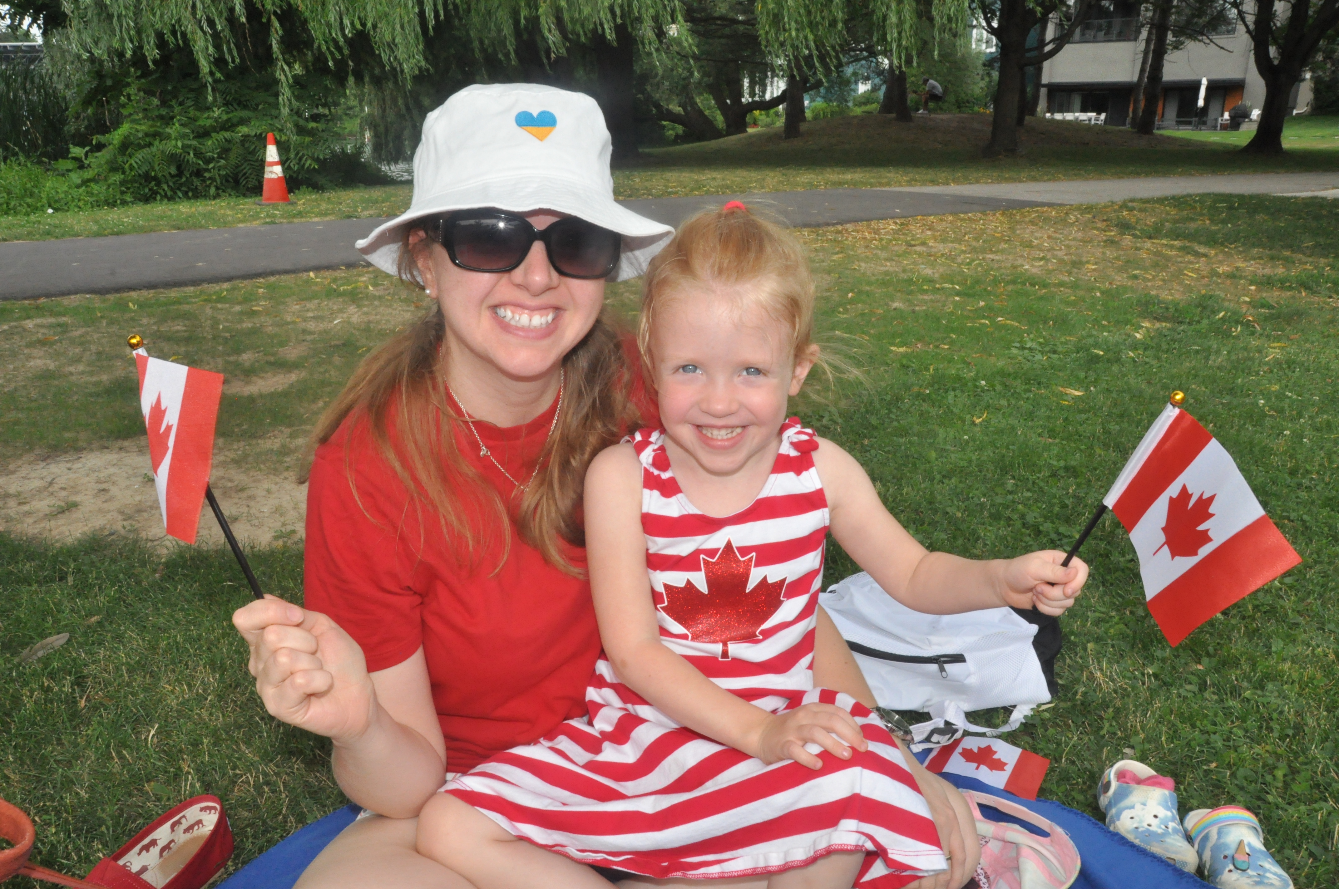 A woman and a small girl sitting on her lap wave Canada Day flags in a parklike setting.