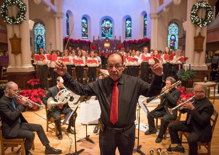 A conductor waves a baton during a Christmas concert, with a choir and musicians performing behind him.