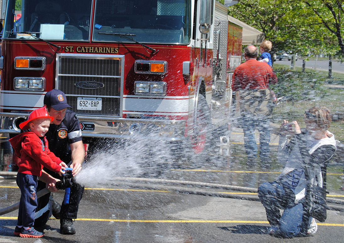 Firefighter showing a child how to use a fire hose while Mom takes a photo.