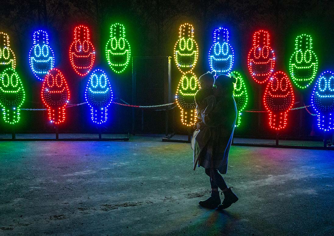 Woman holding a child in front of a Let It Glow lighting display of colourful lightbulb lights.