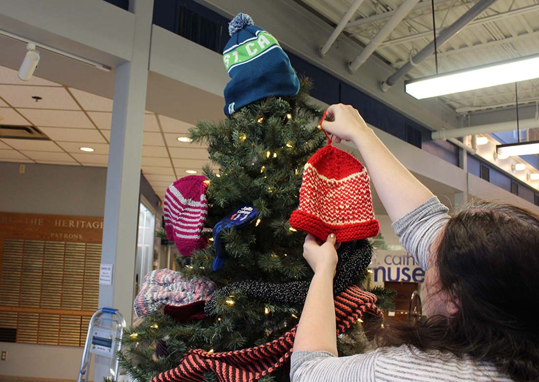 A woman places a red toque winter clothing donation on the Museum's Mitten Tree.