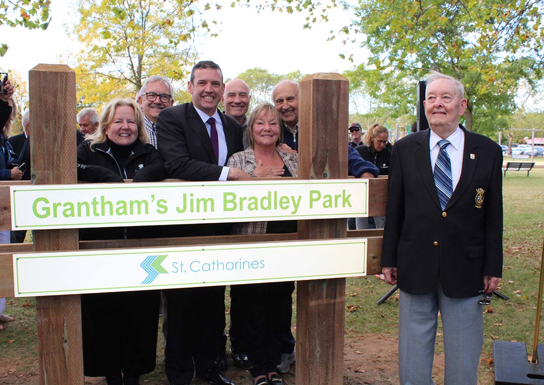 Mayor Mat Siscoe stands at the sign of the renamed Grantham's Jim Bradley Park with the late Regional Chair's brother, Jerry Bradley, far right, and left to right, Grantham Coun. Dawn Dodge, Grantham Coun. Bill Phillips, Port Dalhousie Coun. Bruce Williamson, MPP Jennie Stevens, and St. Andrew's Coun. Joe Kusher on Oct. 8, 2025.