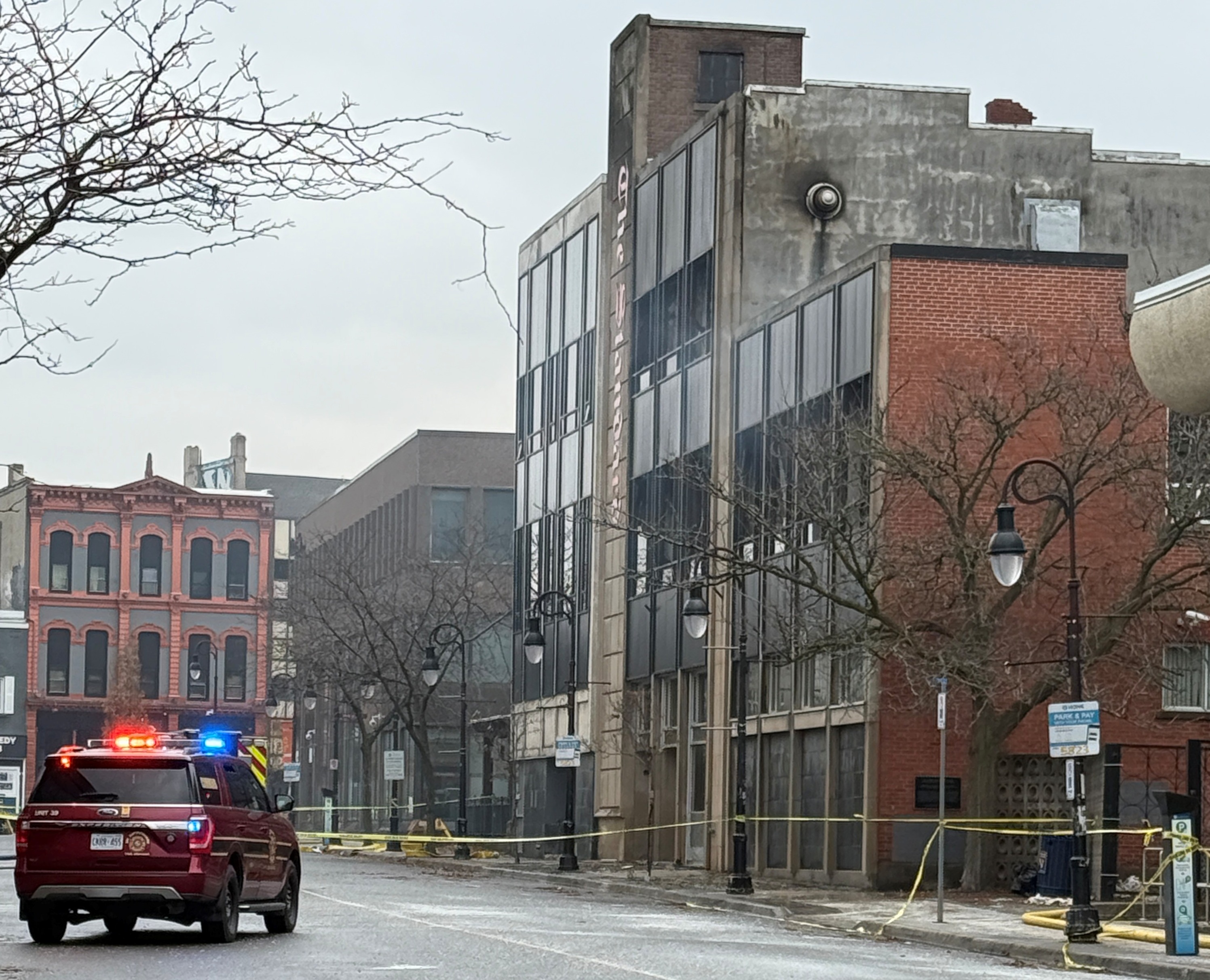 A fire SUV sits on an empty road, with its emergency lights on, in front of a large building with caution tape around it.