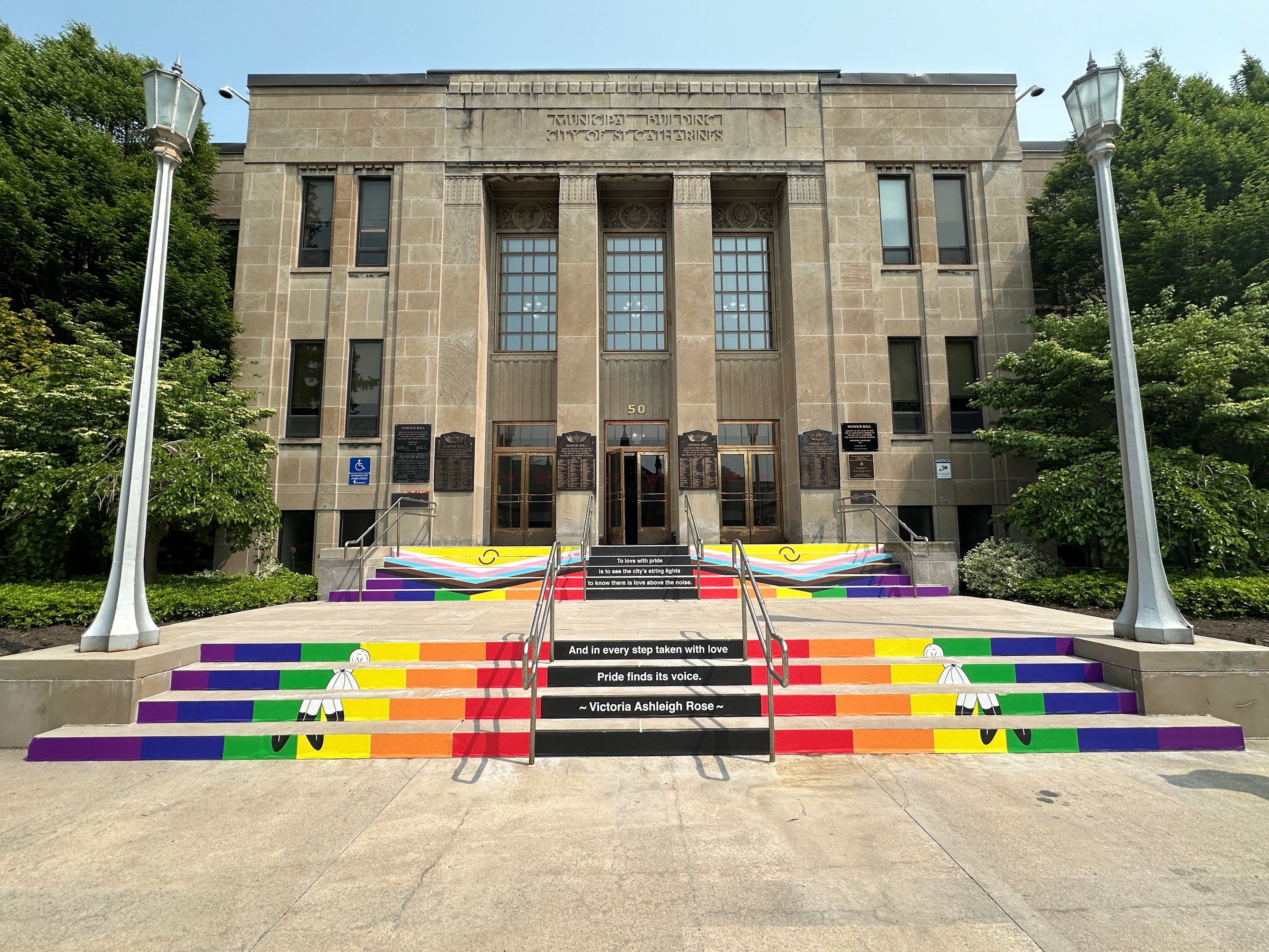 The exterior of St. Catharines City Hall, with the steps leading to the building covered in artwork and a poem for Pride Month