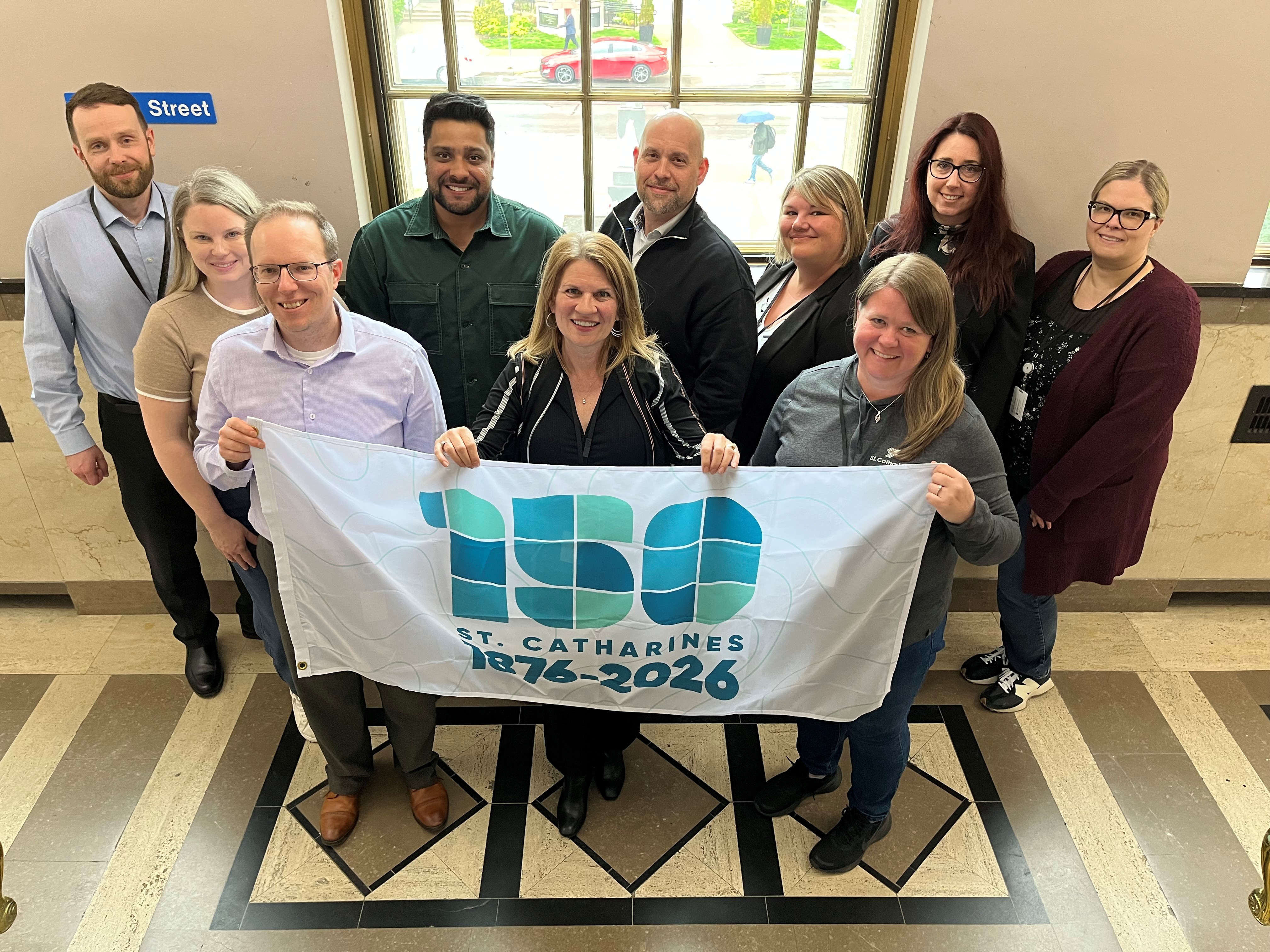 A group of people hold a large flag being used to highlight the 150th anniversary of St. Catharines being incorporated as a City.